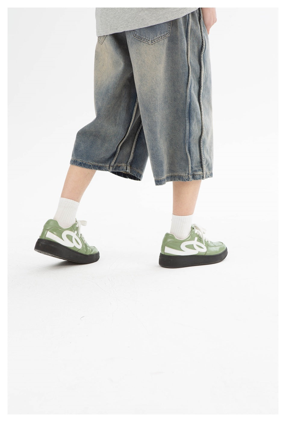 Side-angle detail of loose washed denim shorts with wide legs, worn with white socks and green sneakers.