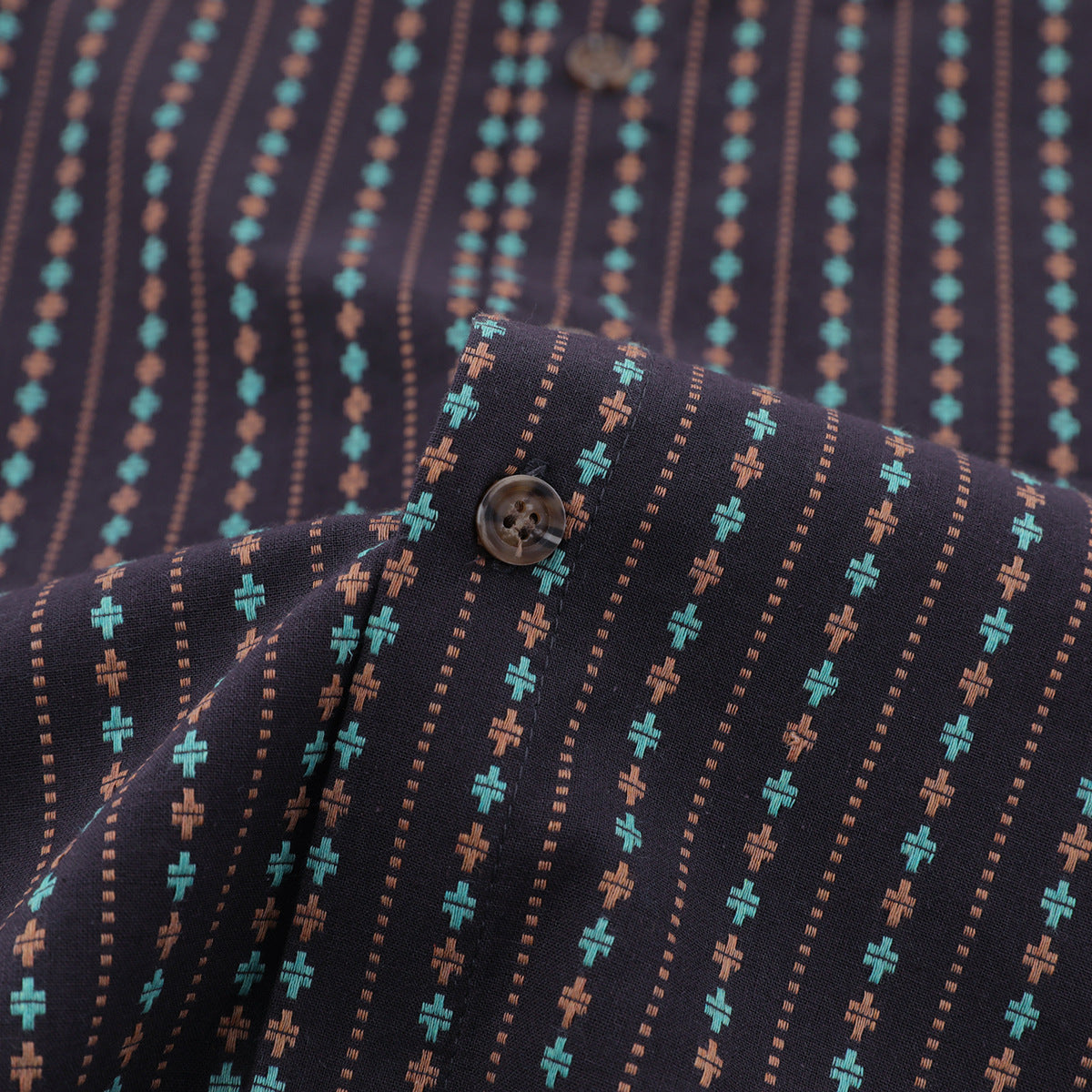 Close-up of a dark shirt with turquoise and rust cross motifs, dotted stripes, and a brown button near the collar.