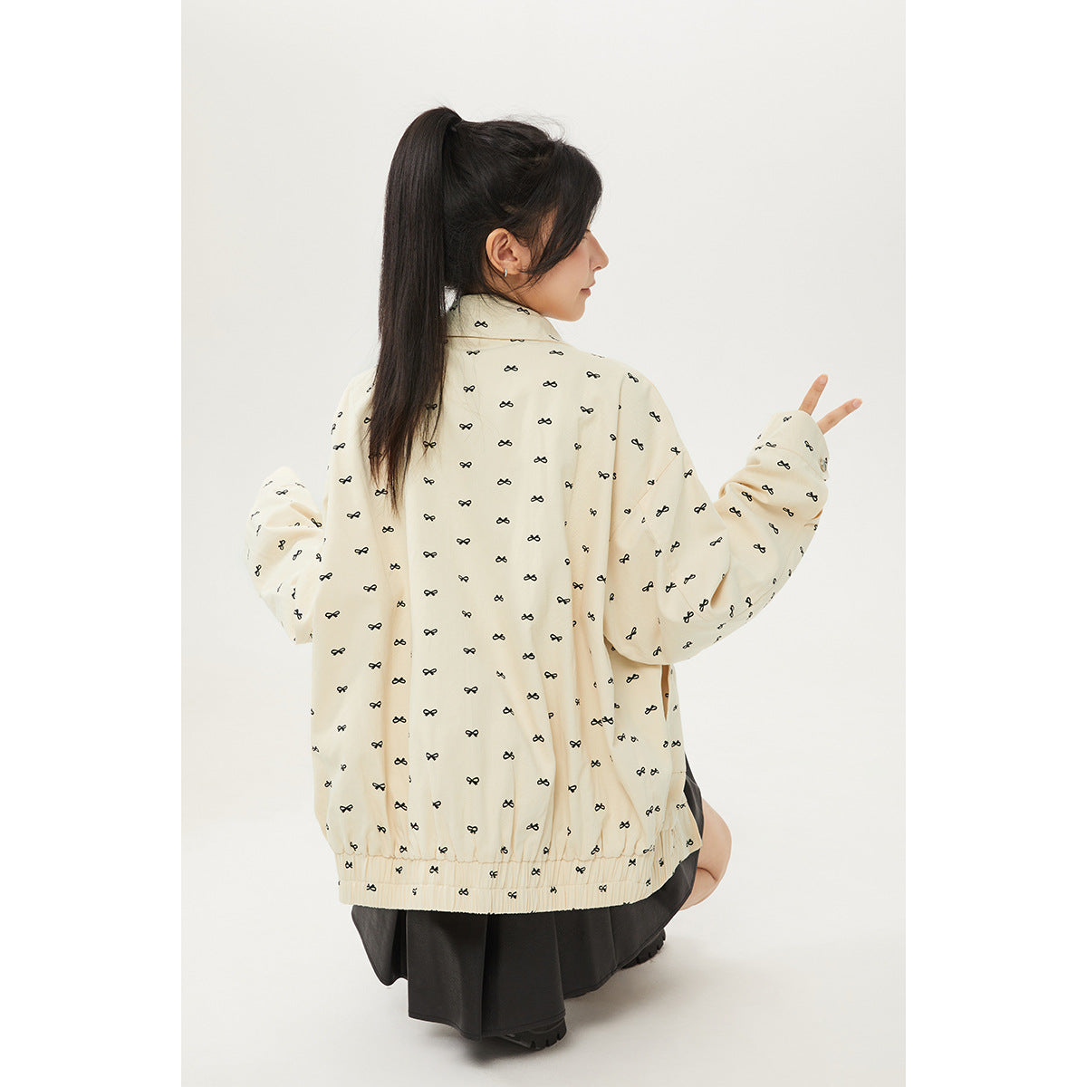 Model showing the back of a cream jacket patterned with small black bows, paired with a black skirt.