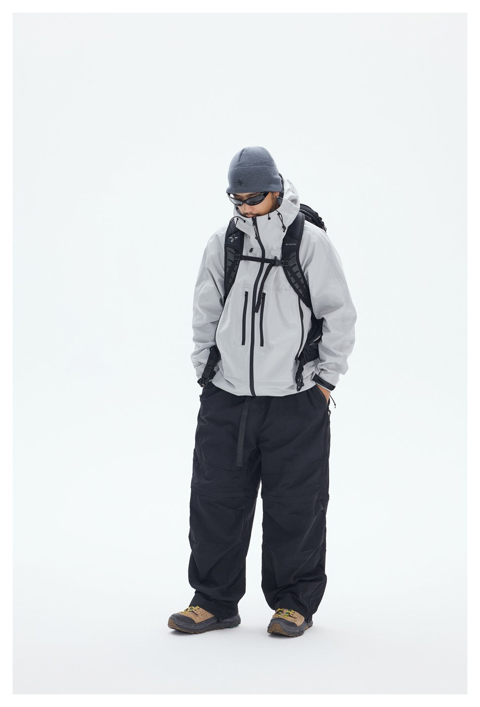 A man wears a light gray hooded jacket, black pants, and a black hiking backpack, shown from the front.