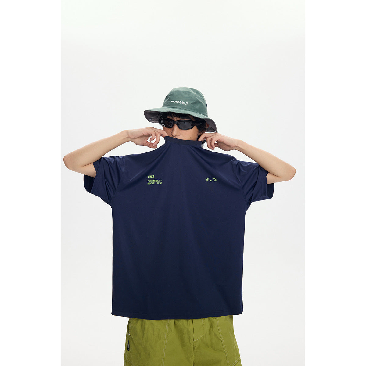 Model holds up an oversized navy T-shirt to show its small lime chest graphics; lime shorts and a green bucket hat are