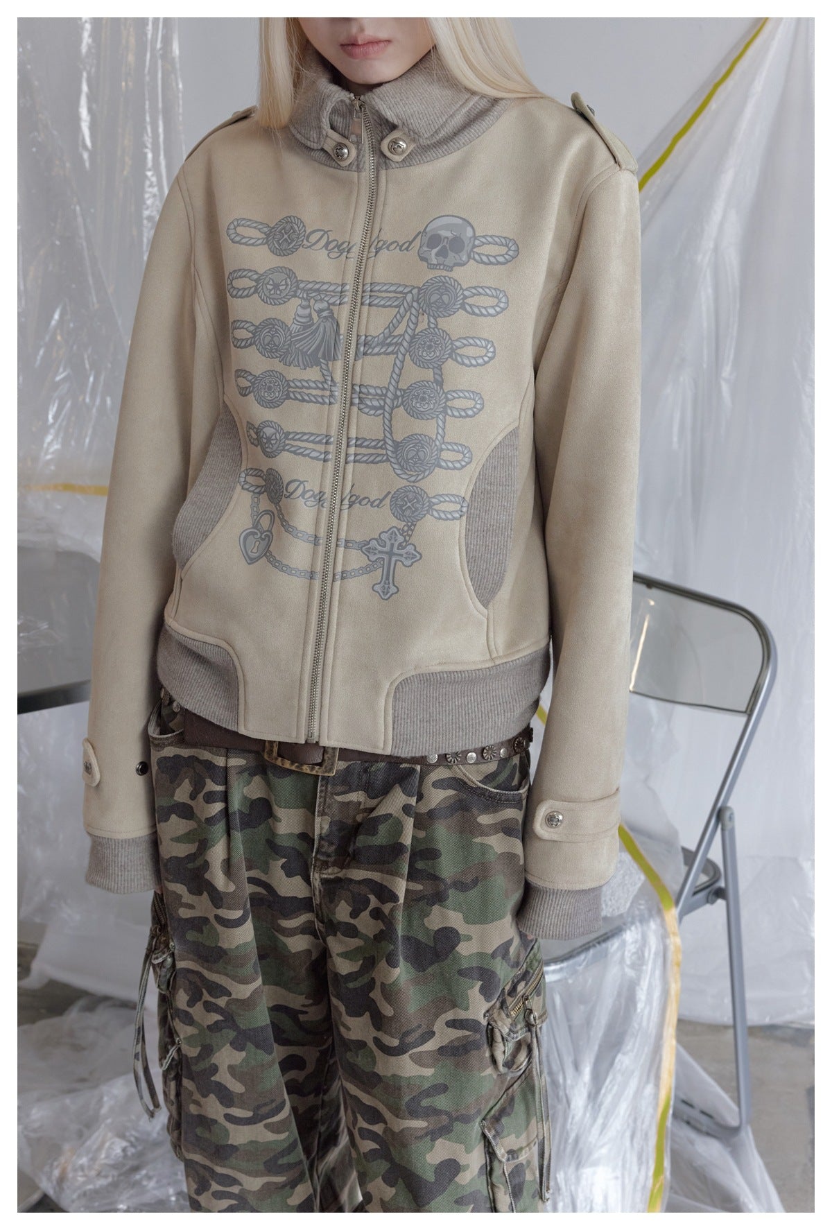 Close-up of a beige zip jacket with a gray rope, skull, and charm graphic, worn with camouflage cargo pants.