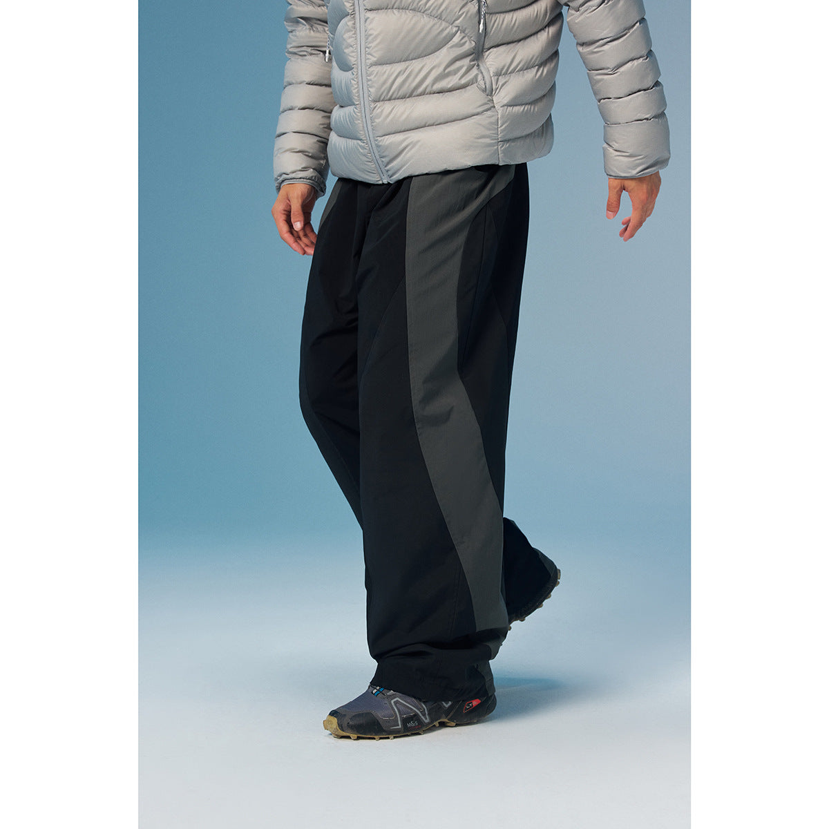 Cropped model wearing loose black trousers with broad charcoal side panels and a silver puffer jacket.
