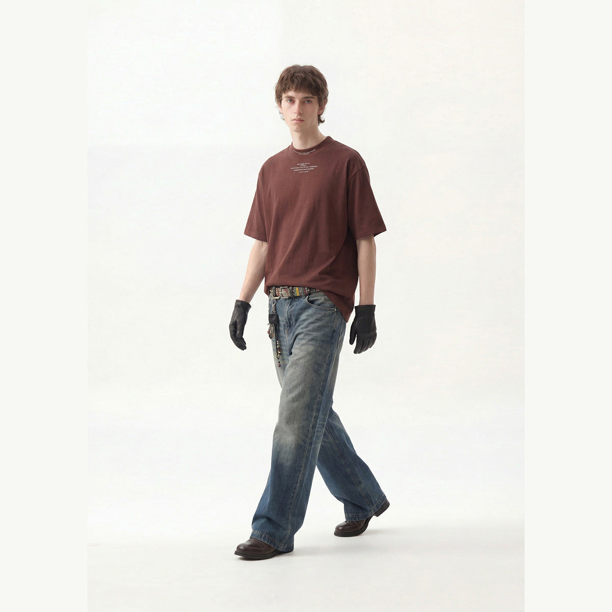 Model wearing a burgundy oversized T-shirt with small chest text, loose blue jeans, black gloves, and dark shoes.