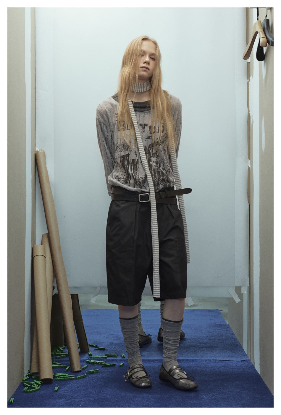Model wearing a gray graphic top, long striped scarf, and black knee-length shorts, shown standing full length.