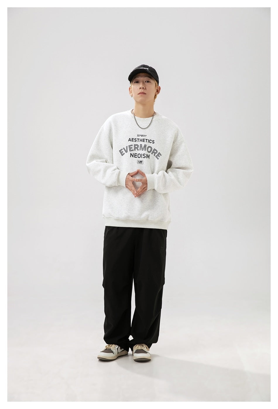 Model wearing a light gray oversized graphic crewneck sweatshirt with black pants, sneakers, and a black cap.