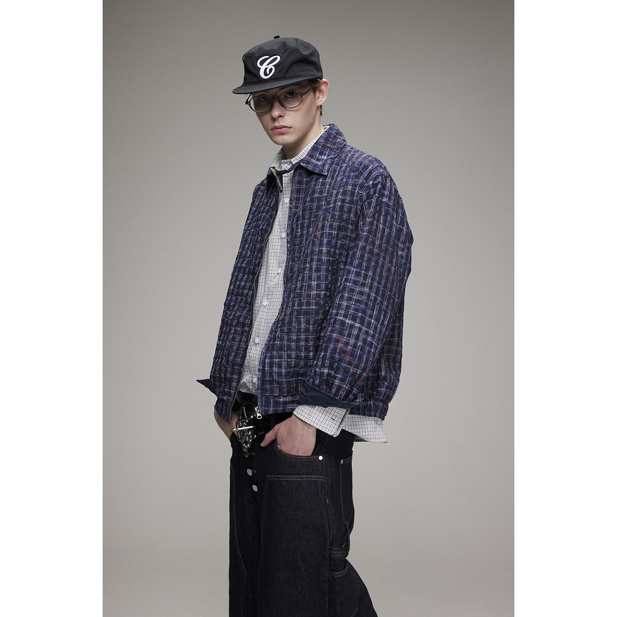 Model wearing a navy plaid jacket over a checked shirt, with dark jeans and a black cap.