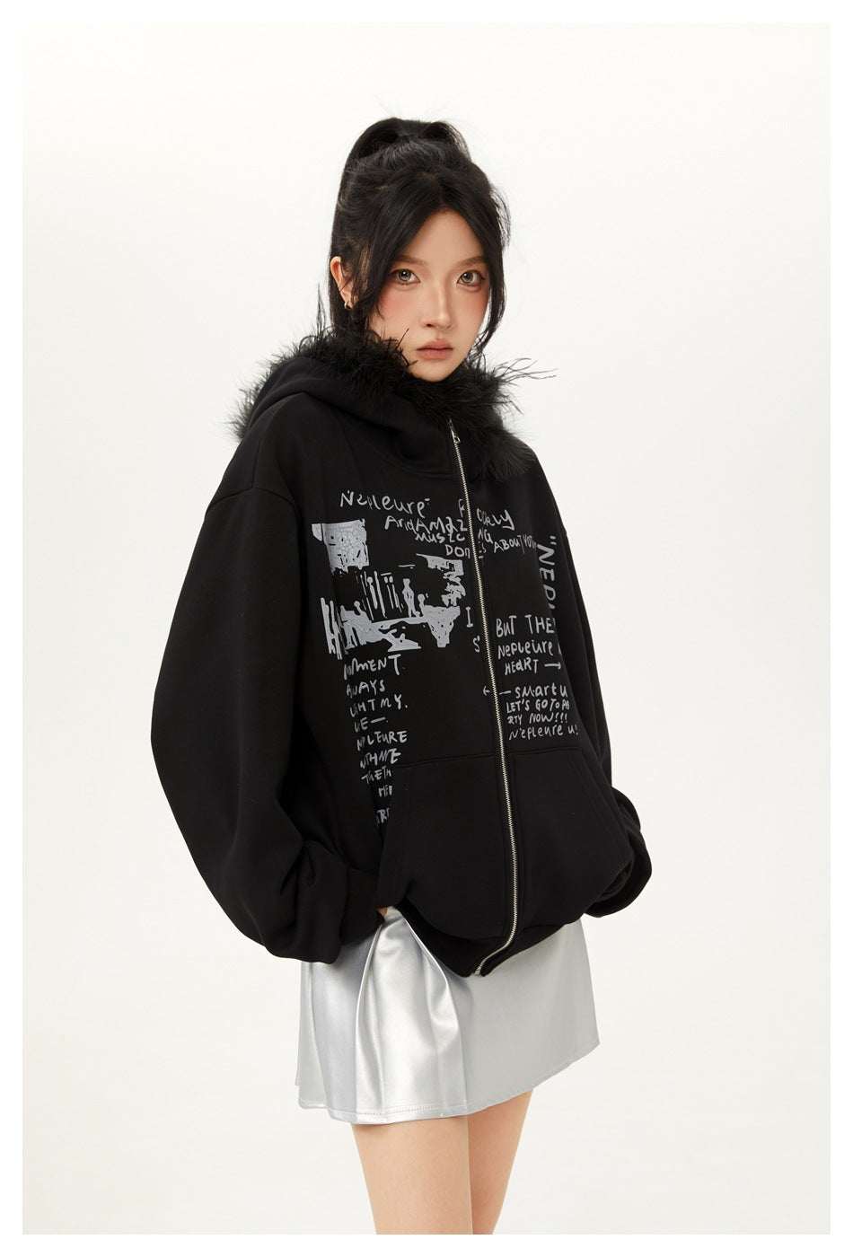 Closer front view of a model in a black fur-trimmed zip hoodie with gray text print and a silver skirt.