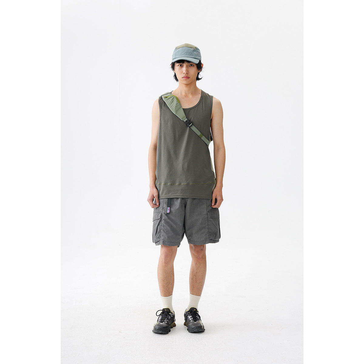 Model wearing a dark olive sleeveless tank and charcoal cargo shorts, with a cap and crossbody strap.