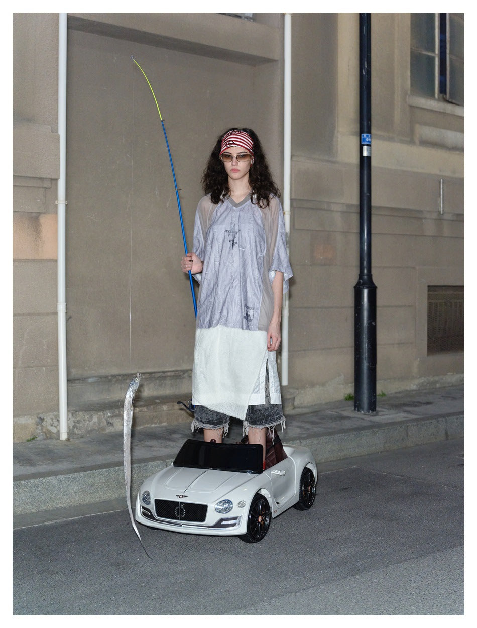 Model standing in a toy car wearing the oversized gray V-neck top with sheer taupe sleeves and a layered white piece