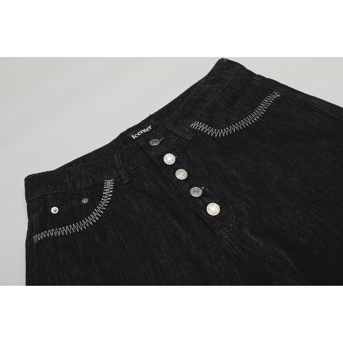 Close-up of black jeans waistband with silver buttons and contrasting white curved pocket stitching.