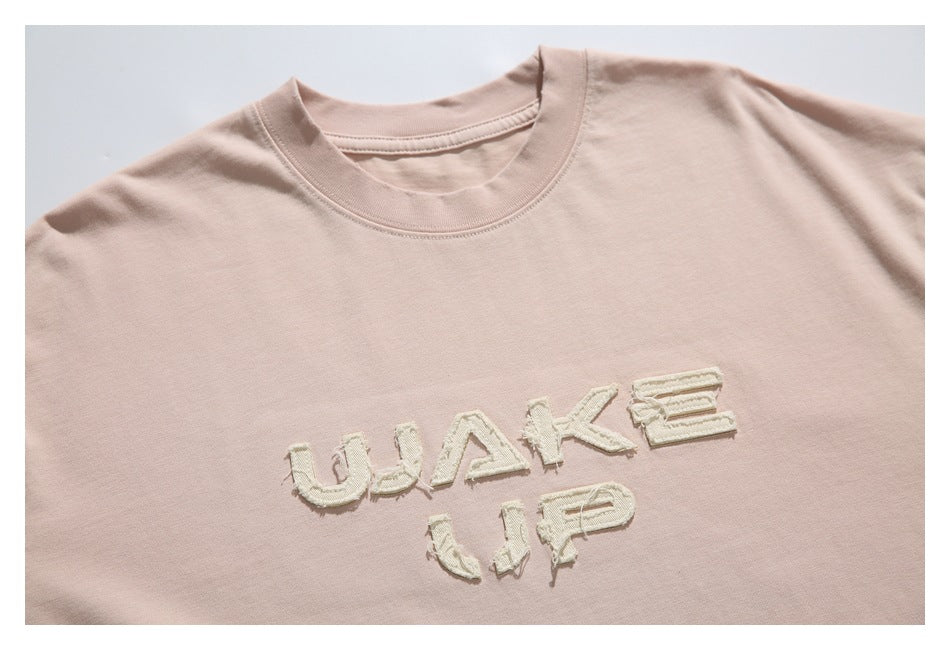 Close view of a pale blush-pink crew-neck T-shirt with cream appliqué lettering reading “WAKE UP” on the chest.