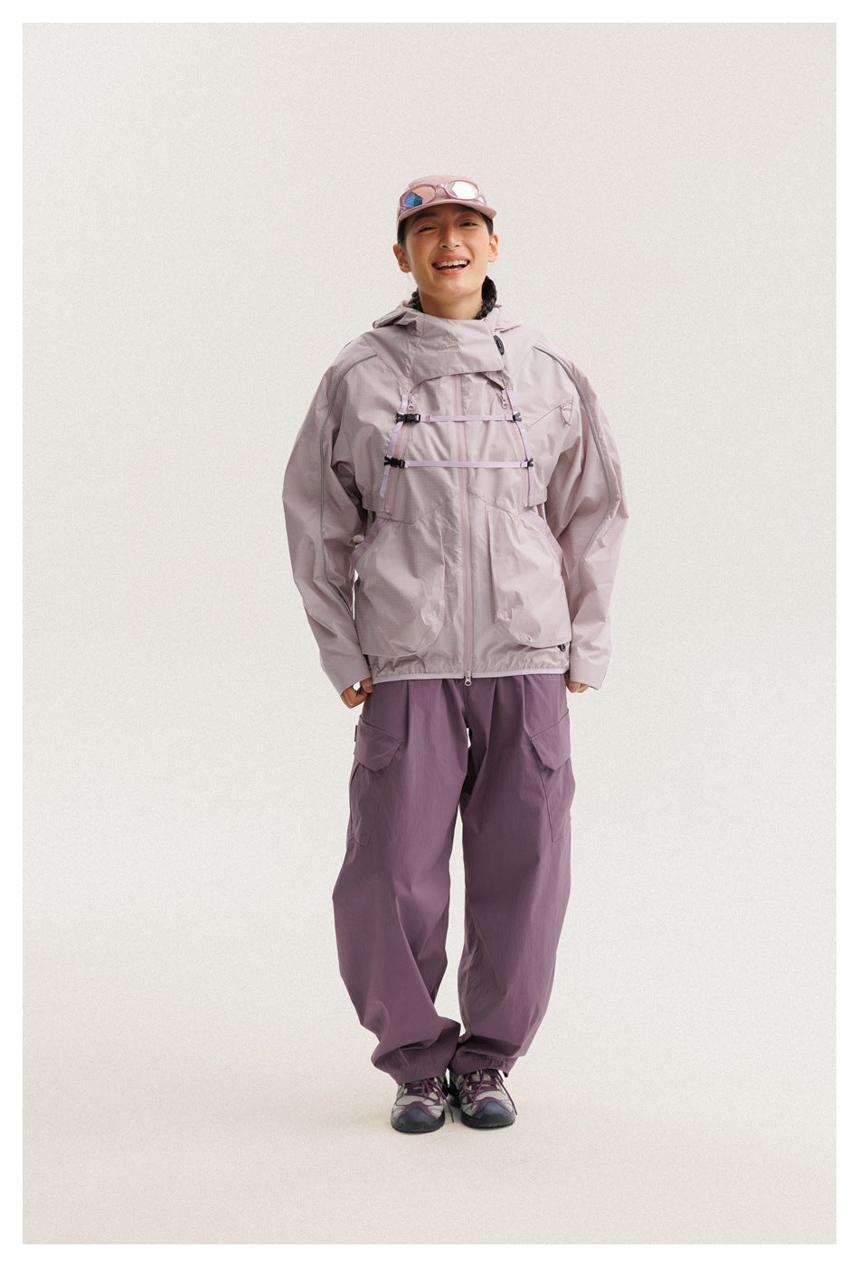 Person wears a pale mauve utility jacket with chest straps and large pockets, plum cargo pants, and a pink cap.