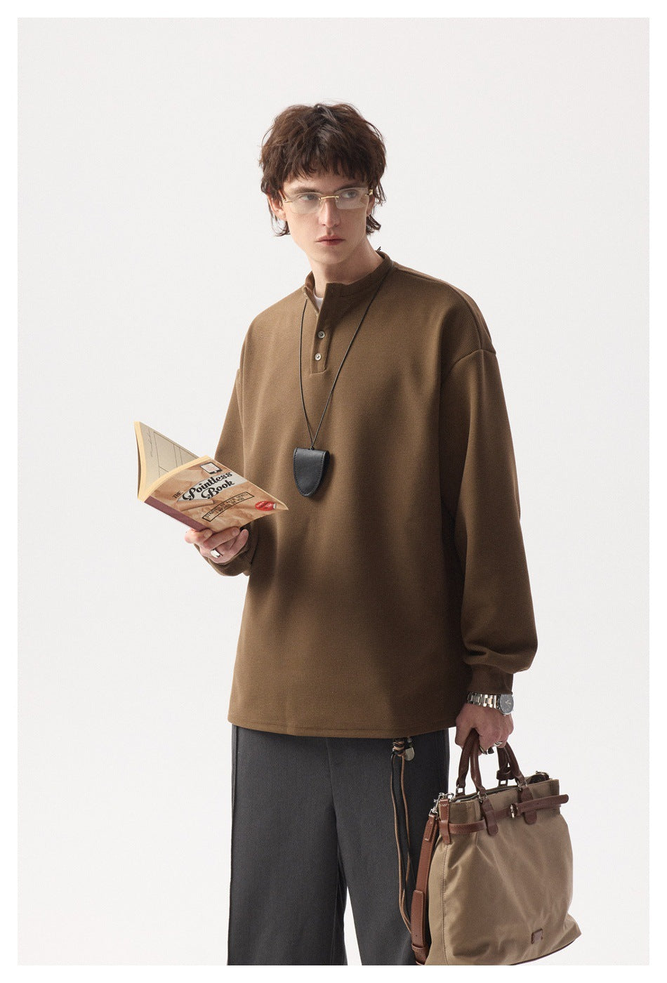 Model wearing brown waffle-knit Henley top with a pendant necklace, reading a book and holding a tote bag