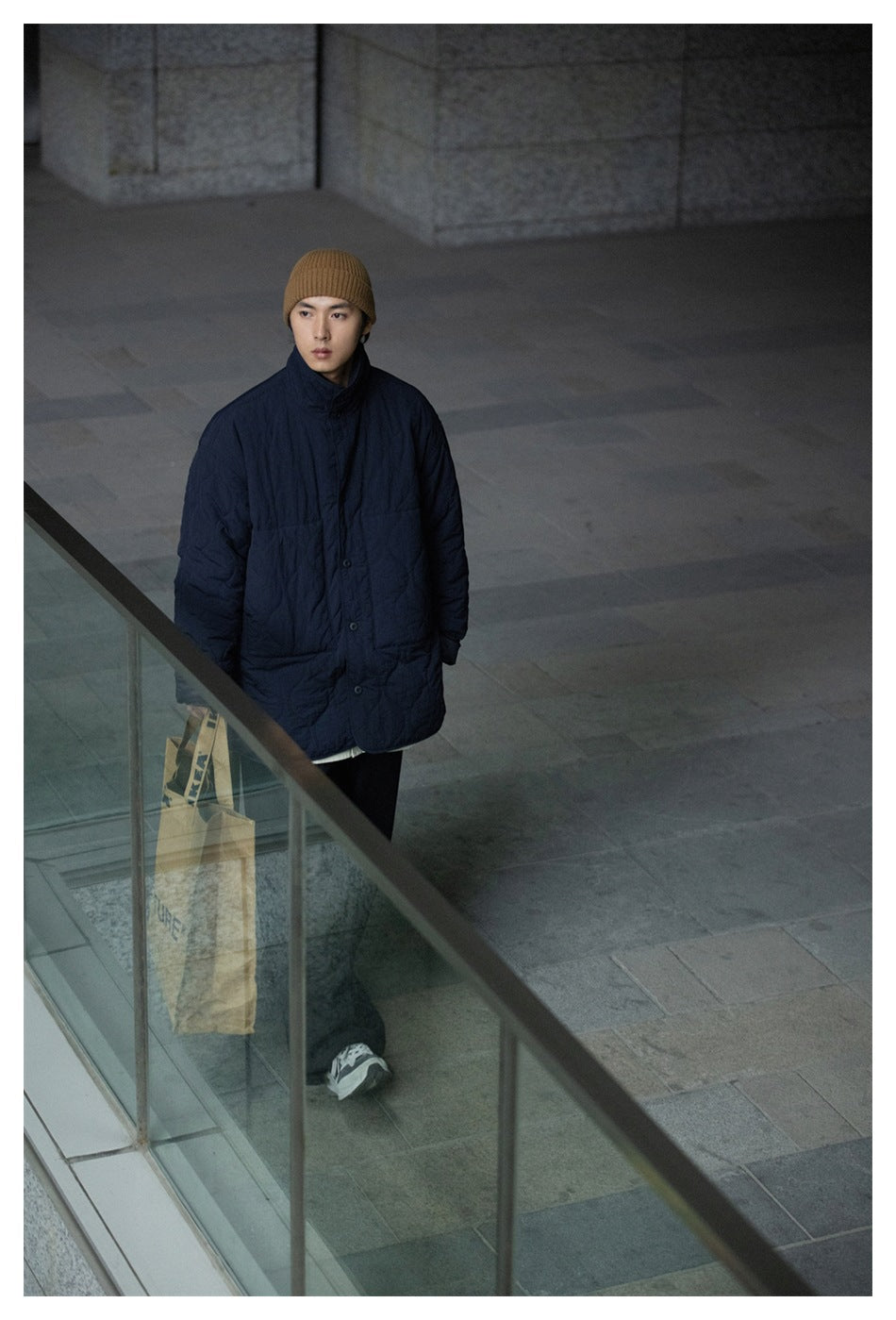 Full-length model wearing a dark navy quilted jacket and brown ribbed beanie, carrying a paper bag.