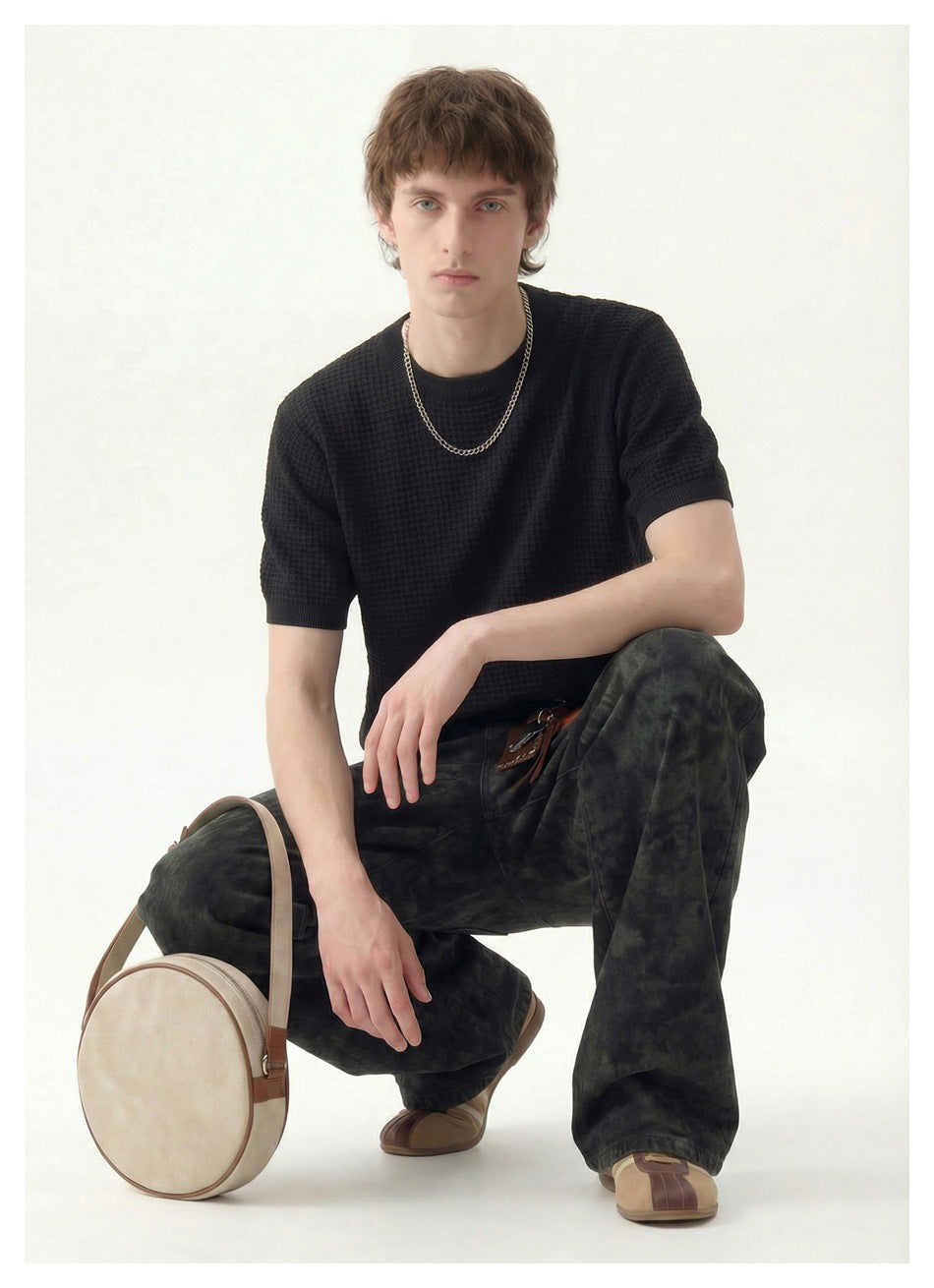 Seated model wearing a black textured short-sleeve crew-neck shirt and dark patterned trousers.