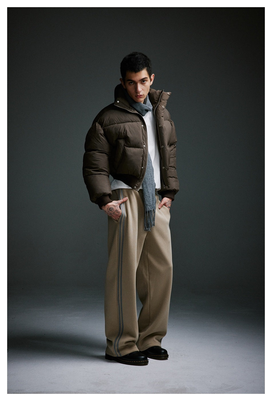 Model wearing a dark brown puffer jacket, gray scarf and wide-leg taupe trousers with side stripes.