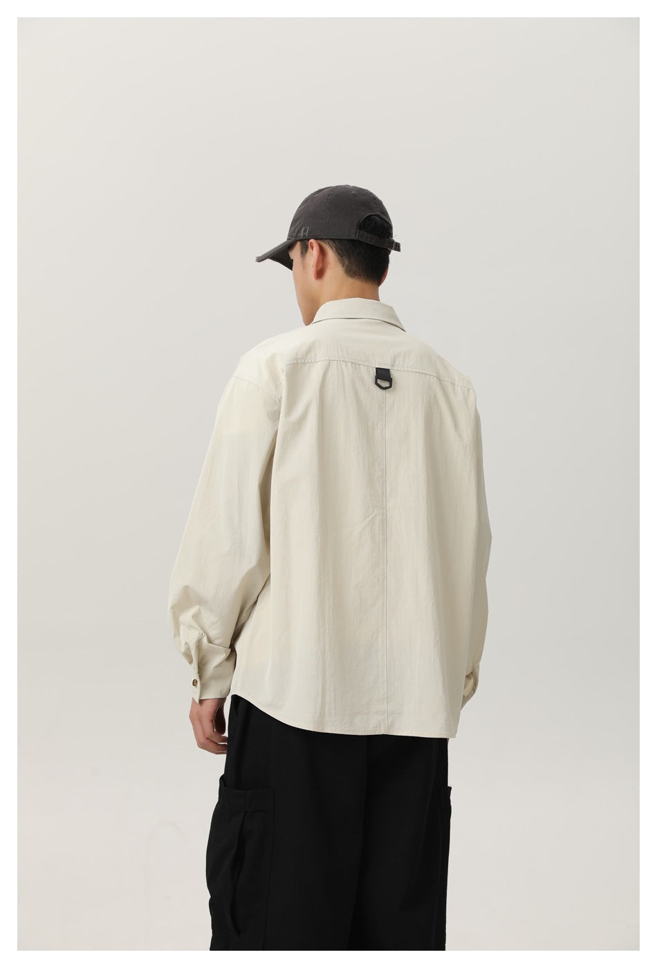 Model wearing a black cap and loose off-white shirt, shown from behind with a small black loop at the upper back.