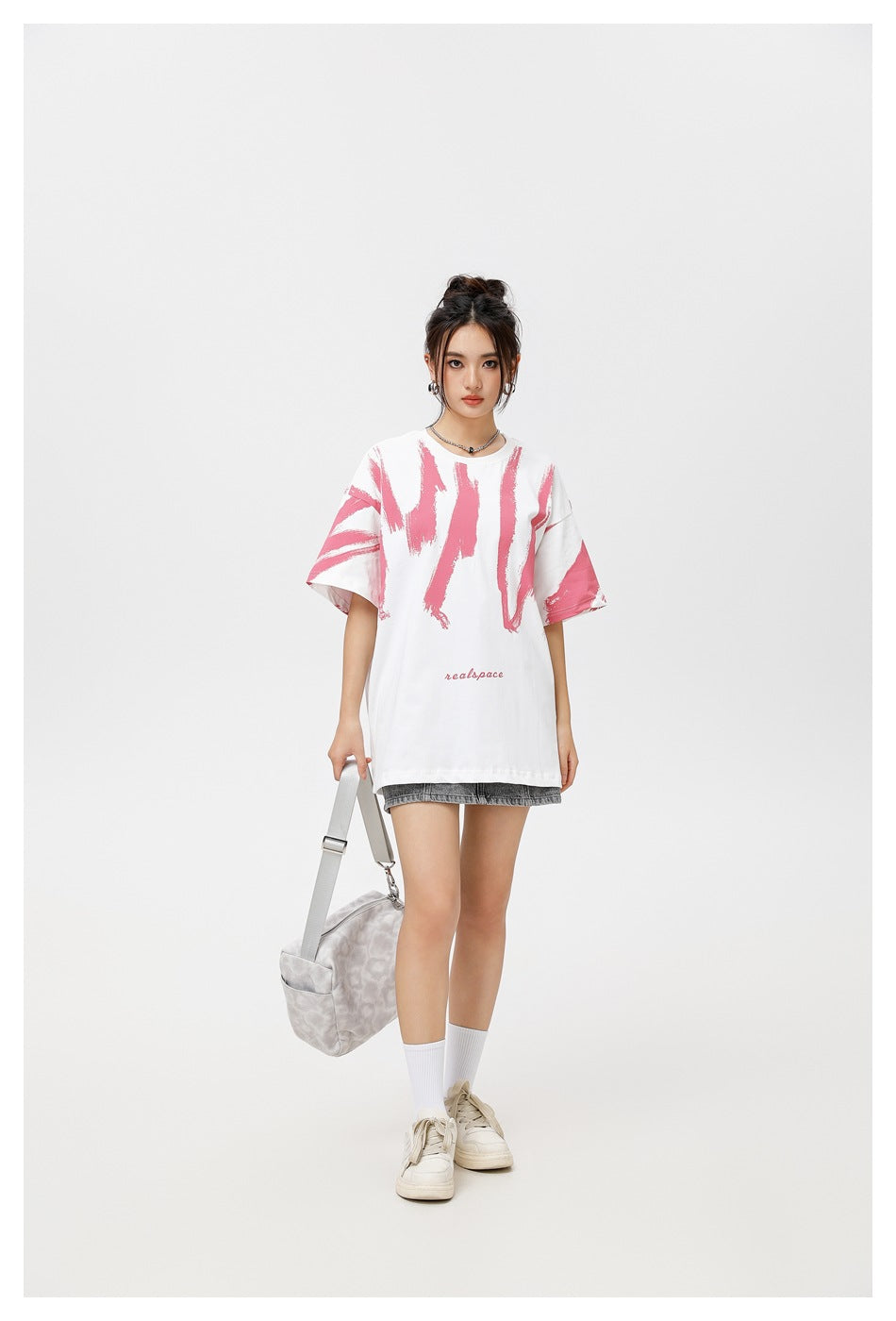 Model wearing an oversized white T-shirt with pink brushstroke graphics and gray shorts, carrying a light shoulder bag.