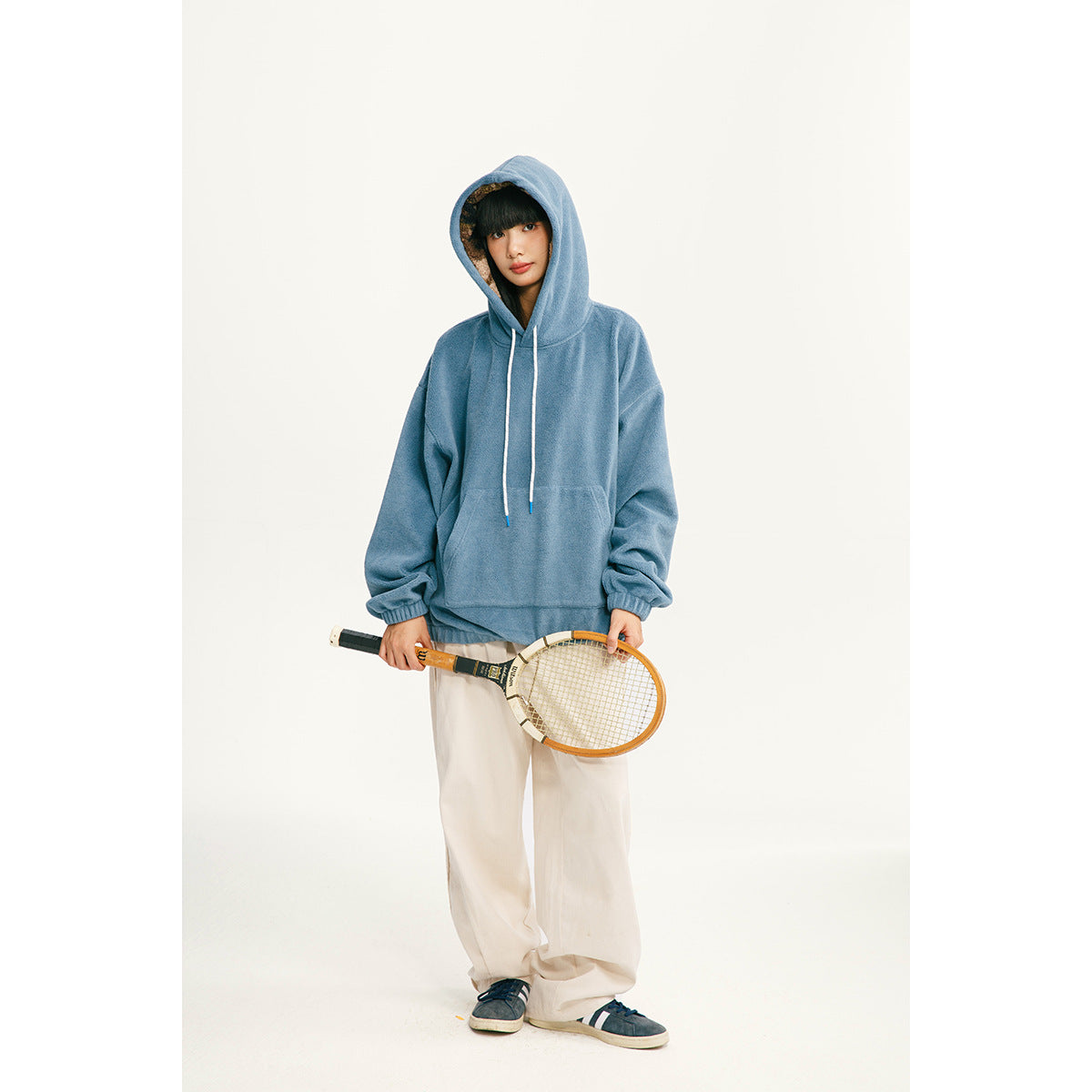 Model wearing a blue pullover hoodie with white drawstrings and a front pocket, paired with loose white pants.