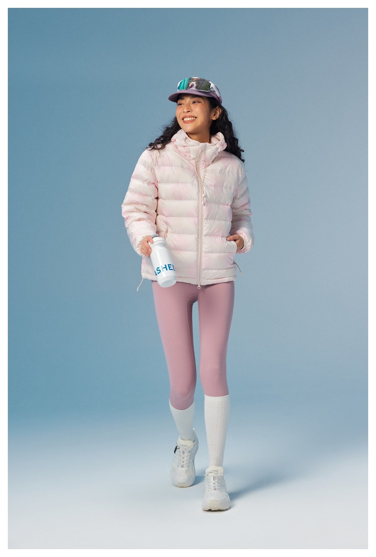 Woman wearing the pink hooded quilted puffer jacket with pink leggings, white socks and sneakers, holding a water bottle