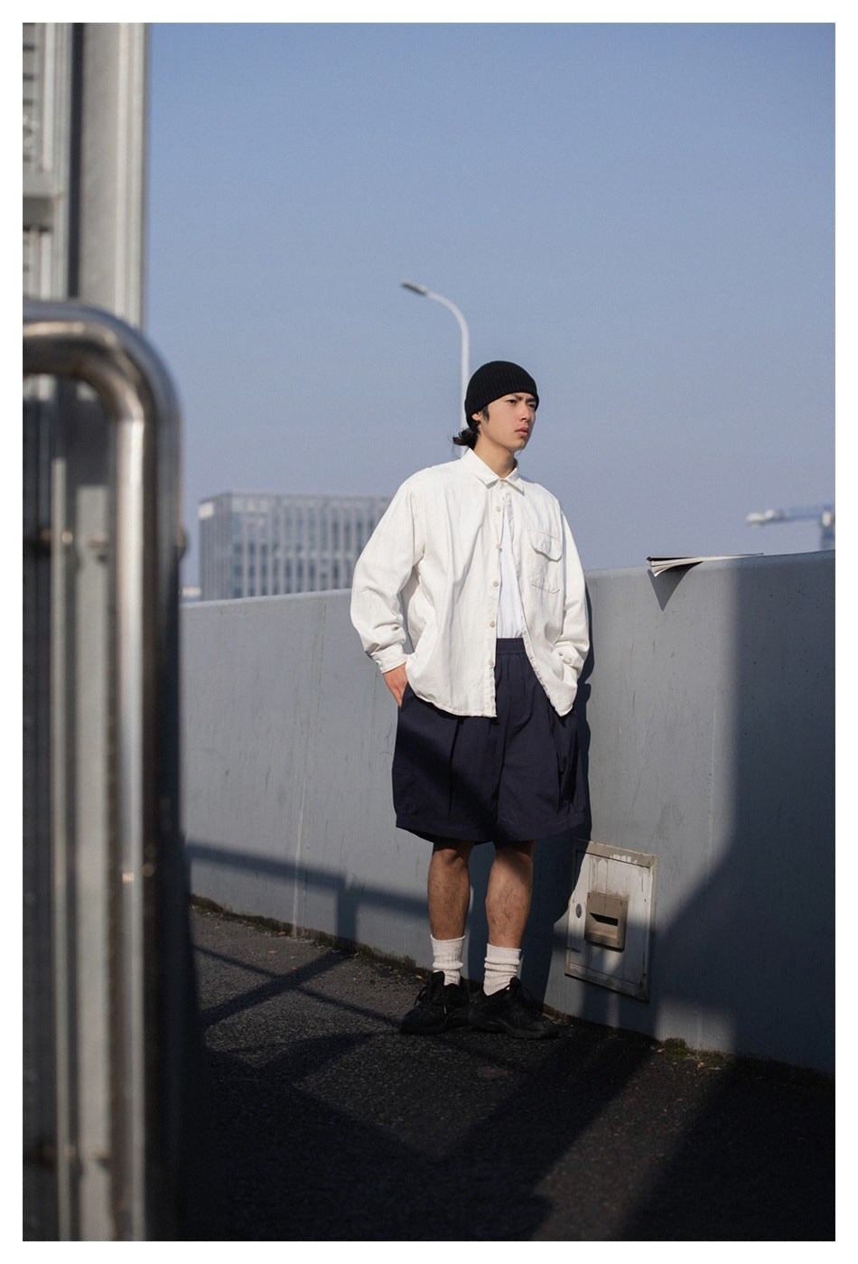 Model wearing a white button-up shirt, navy shorts, black beanie, white socks and black sneakers outdoors.