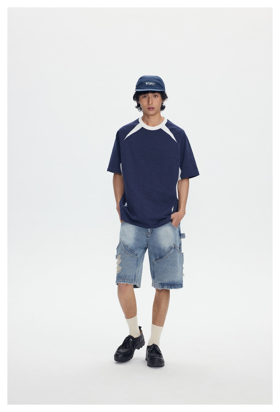 Model wearing navy color-block raglan T-shirt with white trim, denim shorts, cap and black shoes, front view