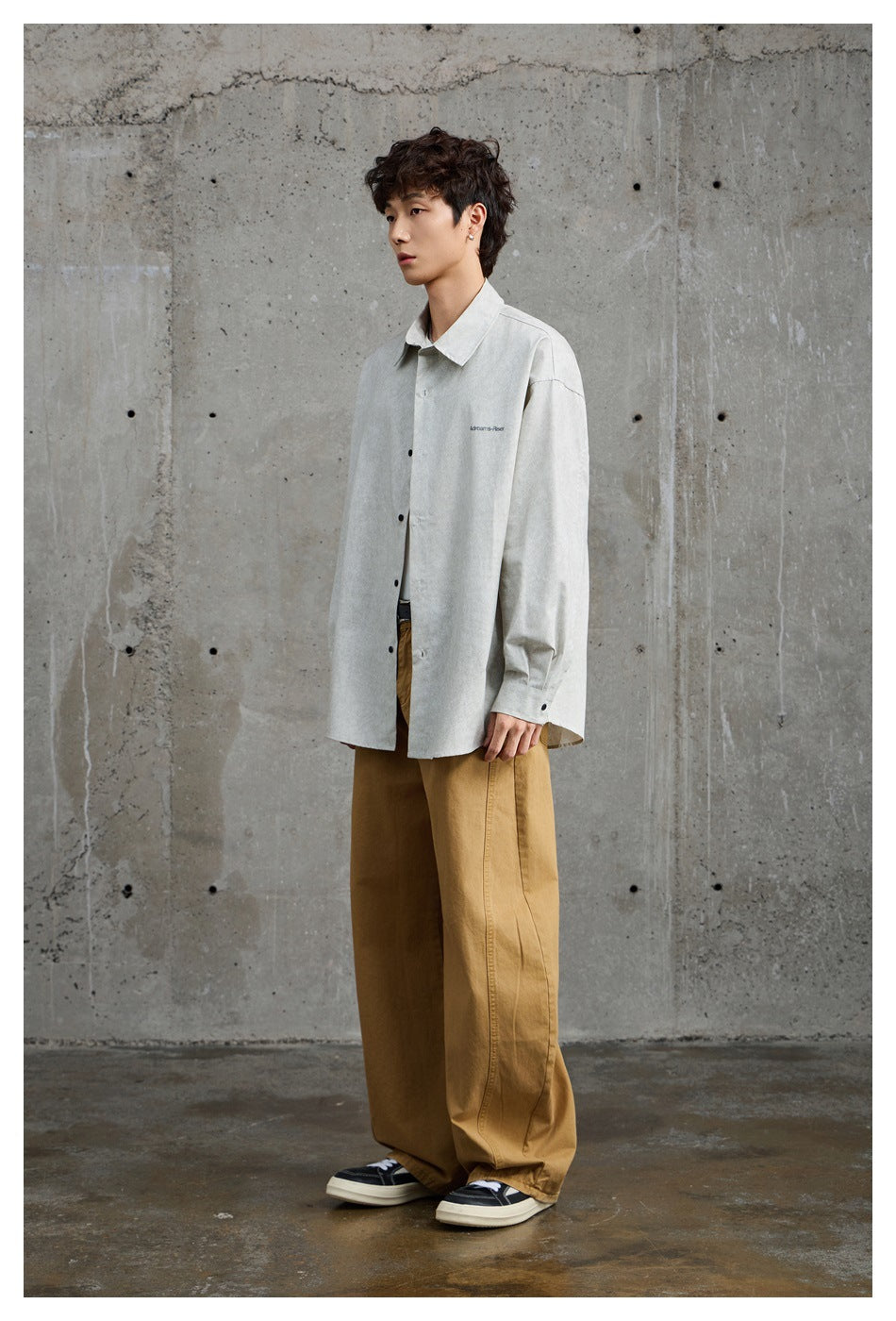 Model wearing an oversized light gray button-up shirt with black buttons and small chest lettering, paired with tan wide-leg