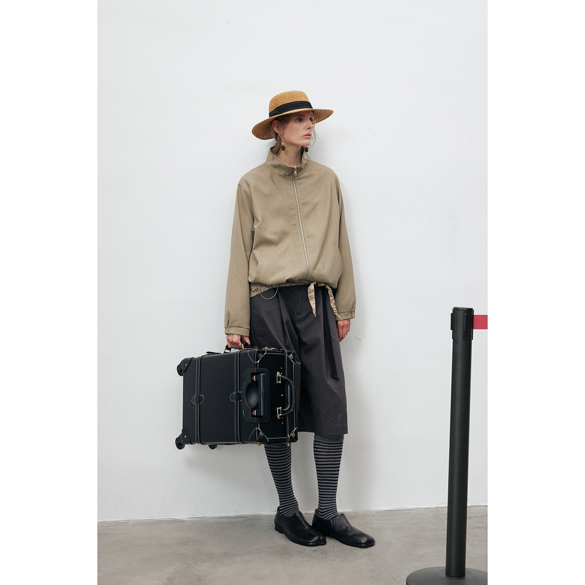 Model wearing a taupe zip-front jacket with a stand collar, dark skirt, and striped socks, carrying a black suitcase.