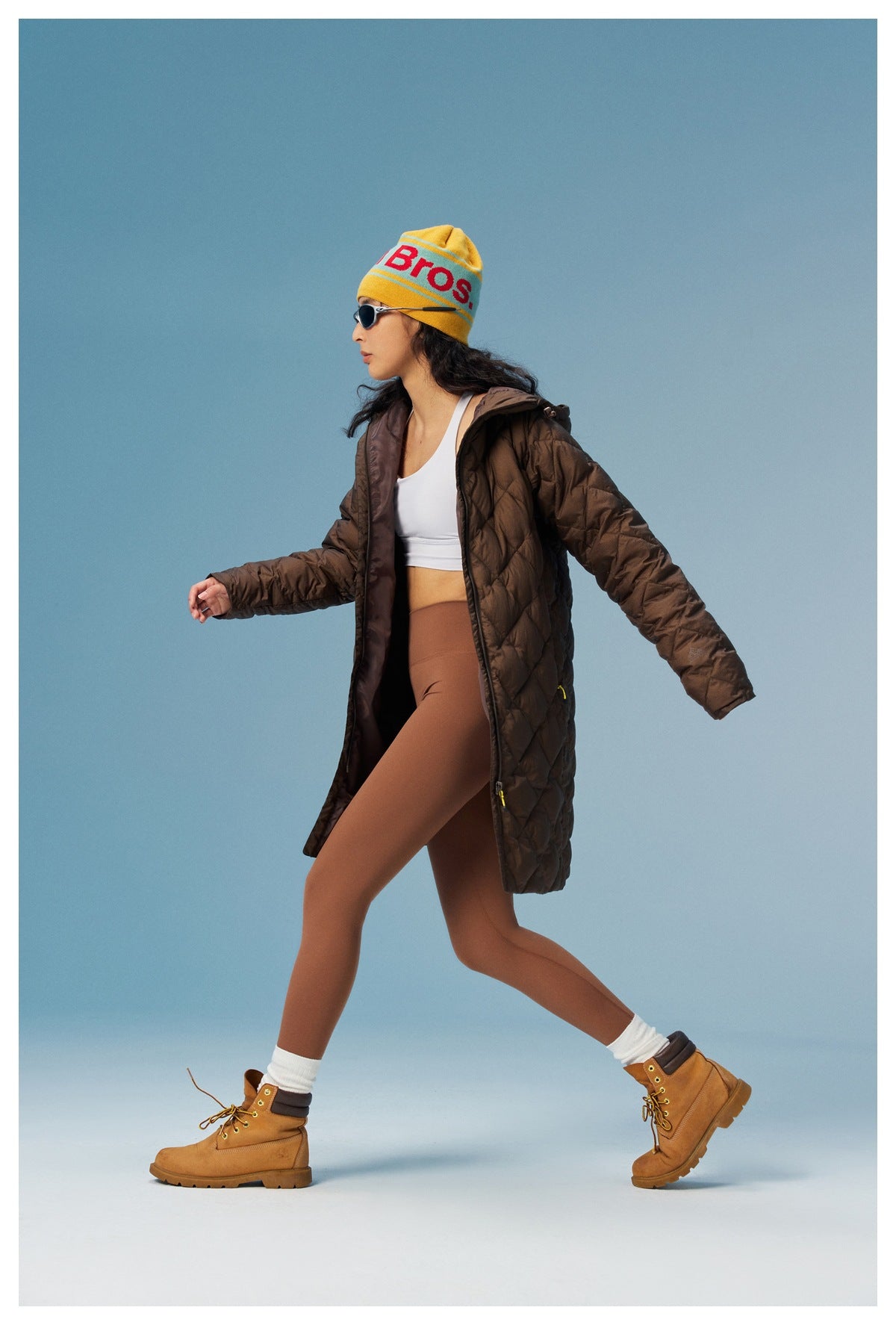 Model walking in an open brown quilted hooded coat over a white top and brown leggings.