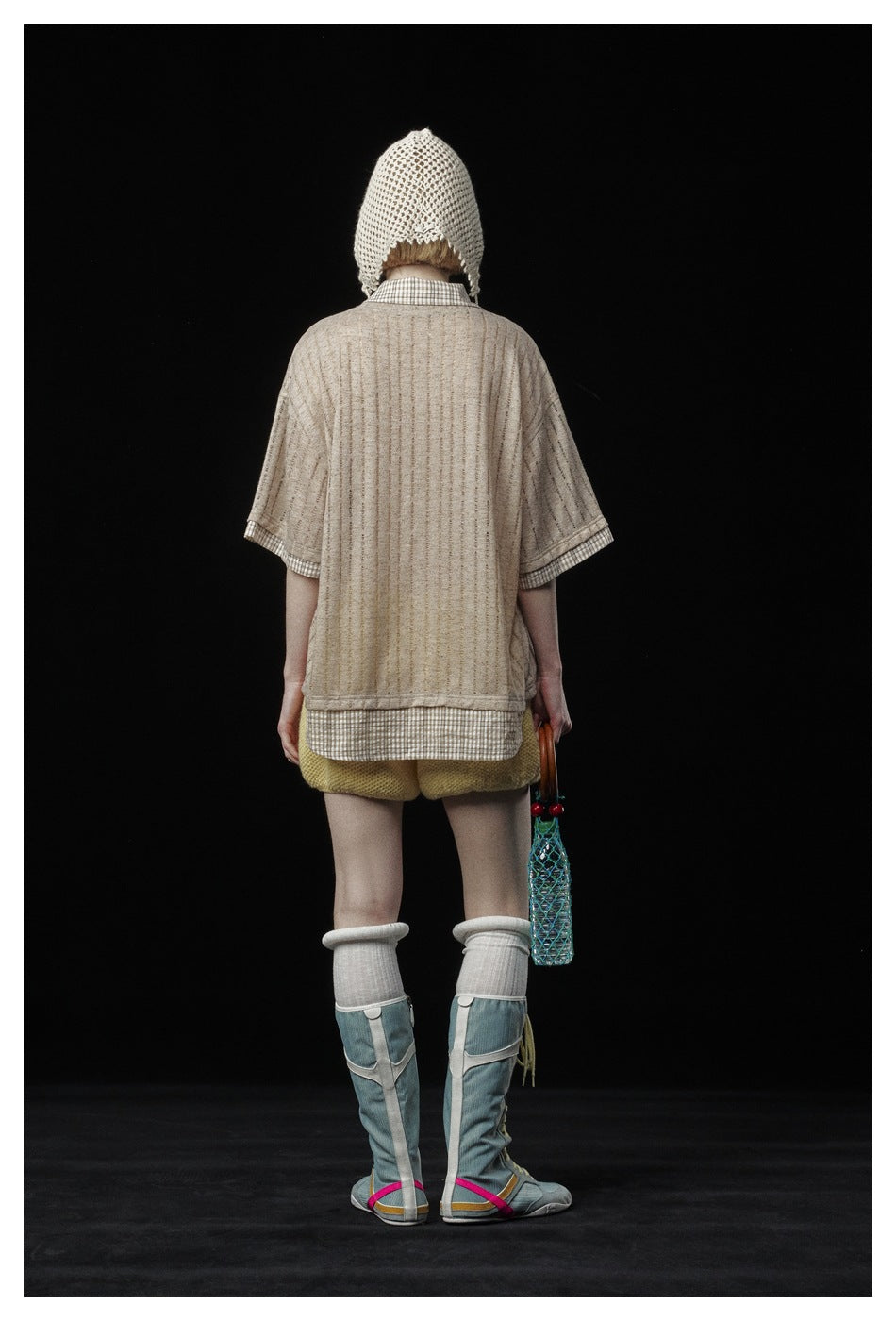 Back view of a model wearing a beige striped tee over a checked shirt with yellow shorts, a cream crochet bonnet, and tall