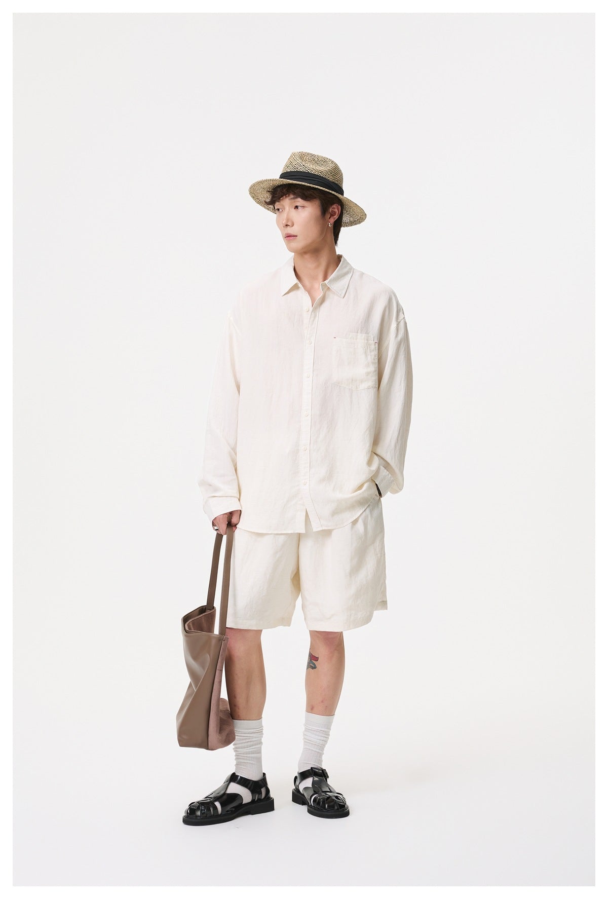 Model wearing an ivory button-up shirt and matching shorts with white socks, black sandals, a straw hat, and brown tote.