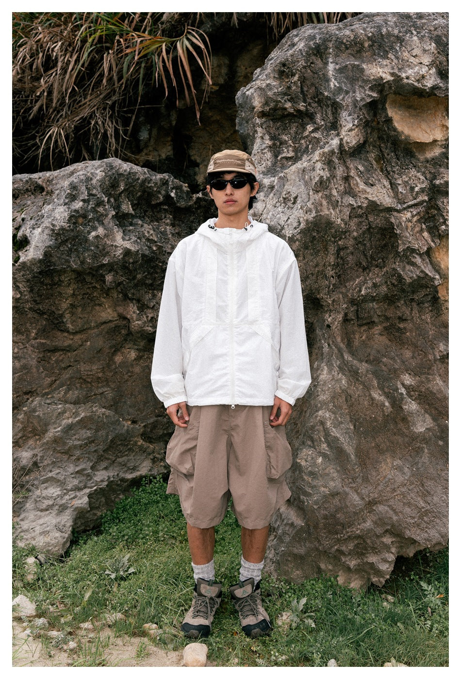 A man stands among rocks wearing a white hooded zip jacket and taupe cargo shorts.