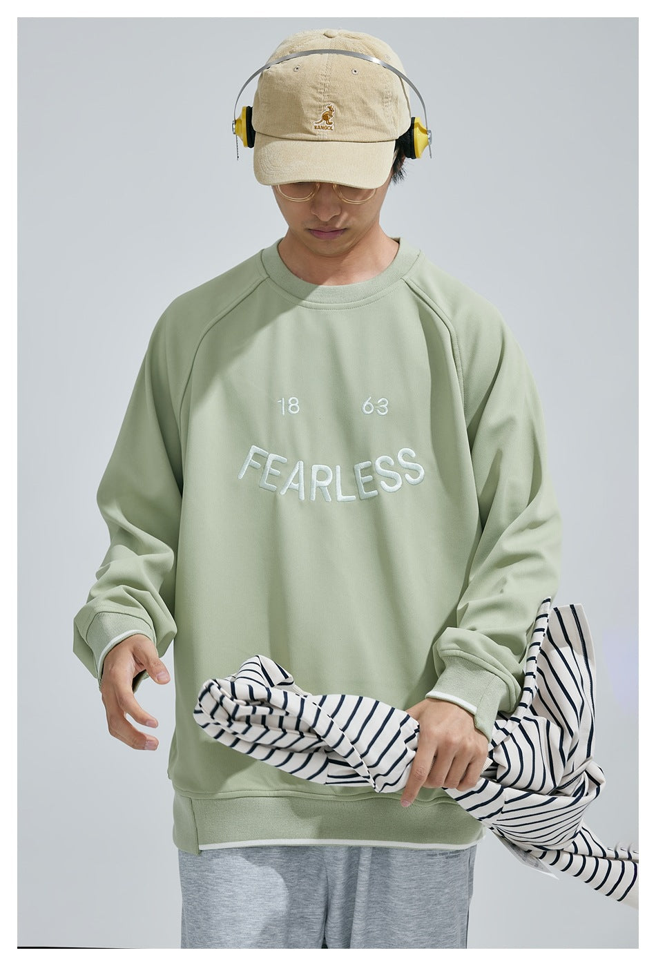 Front view of a model wearing a sage-green raglan sweatshirt with white “18 63 FEARLESS” embroidery.