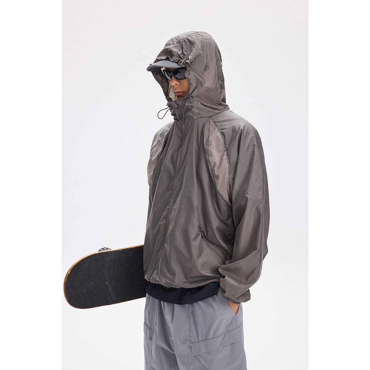 Model in a dark gray hooded zip jacket and gray shorts, holding a skateboard.