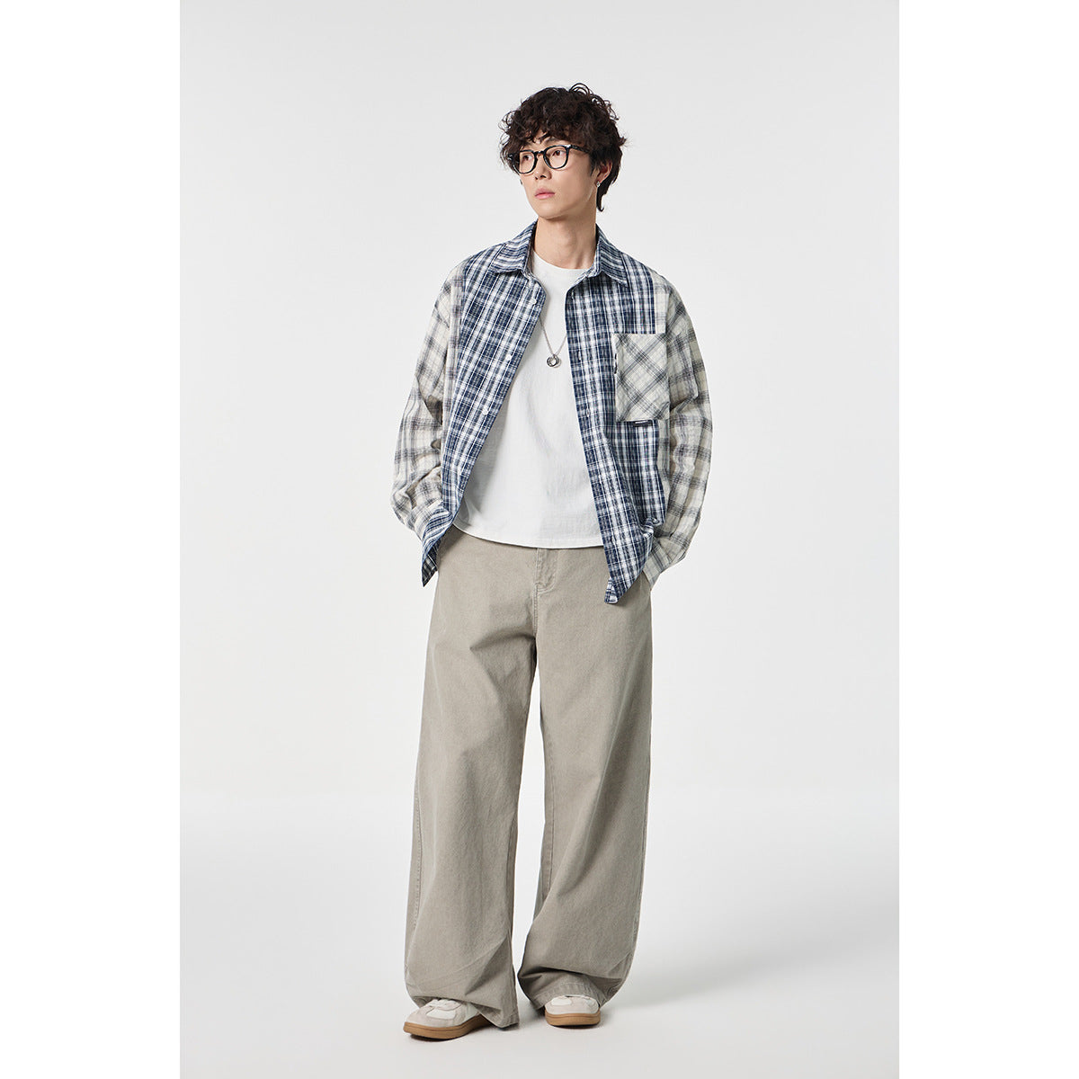 Model wearing an open blue-and-gray plaid shirt with a chest pocket over a white tee and loose gray trousers.