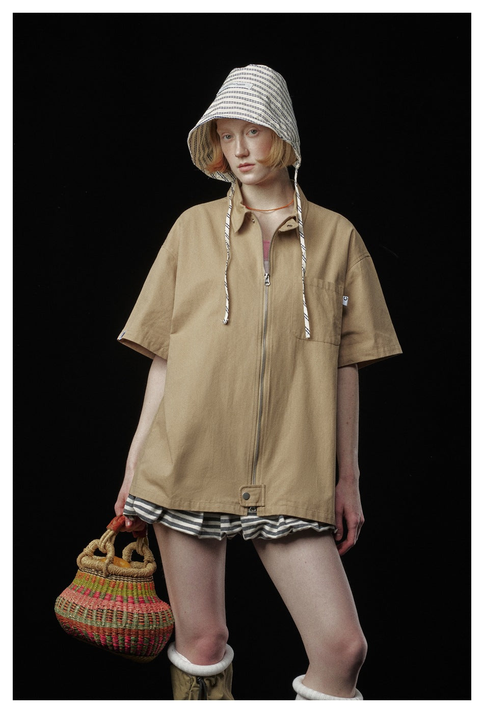 Model wearing a tan short-sleeve zip jacket with a chest pocket, striped bucket hat and shorts.
