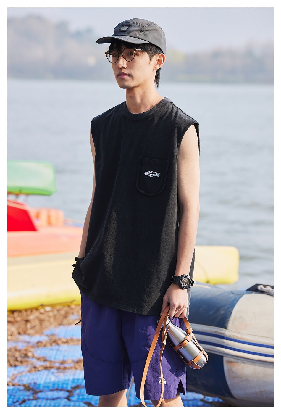 Model in a black sleeveless pocket shirt and purple shorts, shown from the front by the water.