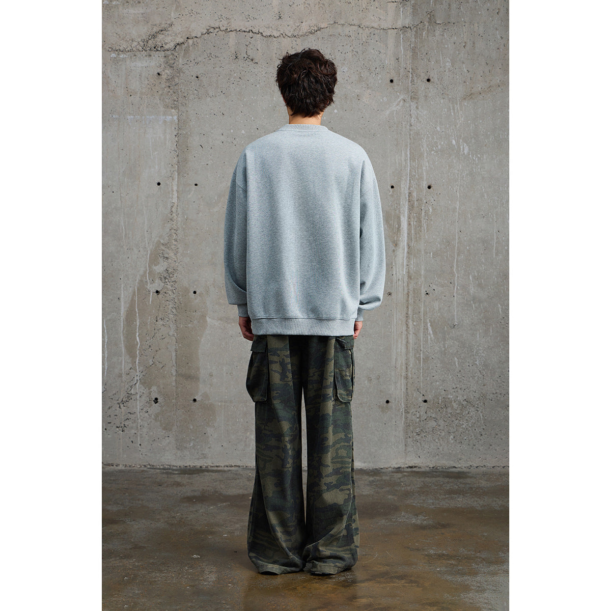 Rear view of a model wearing an oversized light gray crewneck sweatshirt and loose dark camouflage cargo pants.