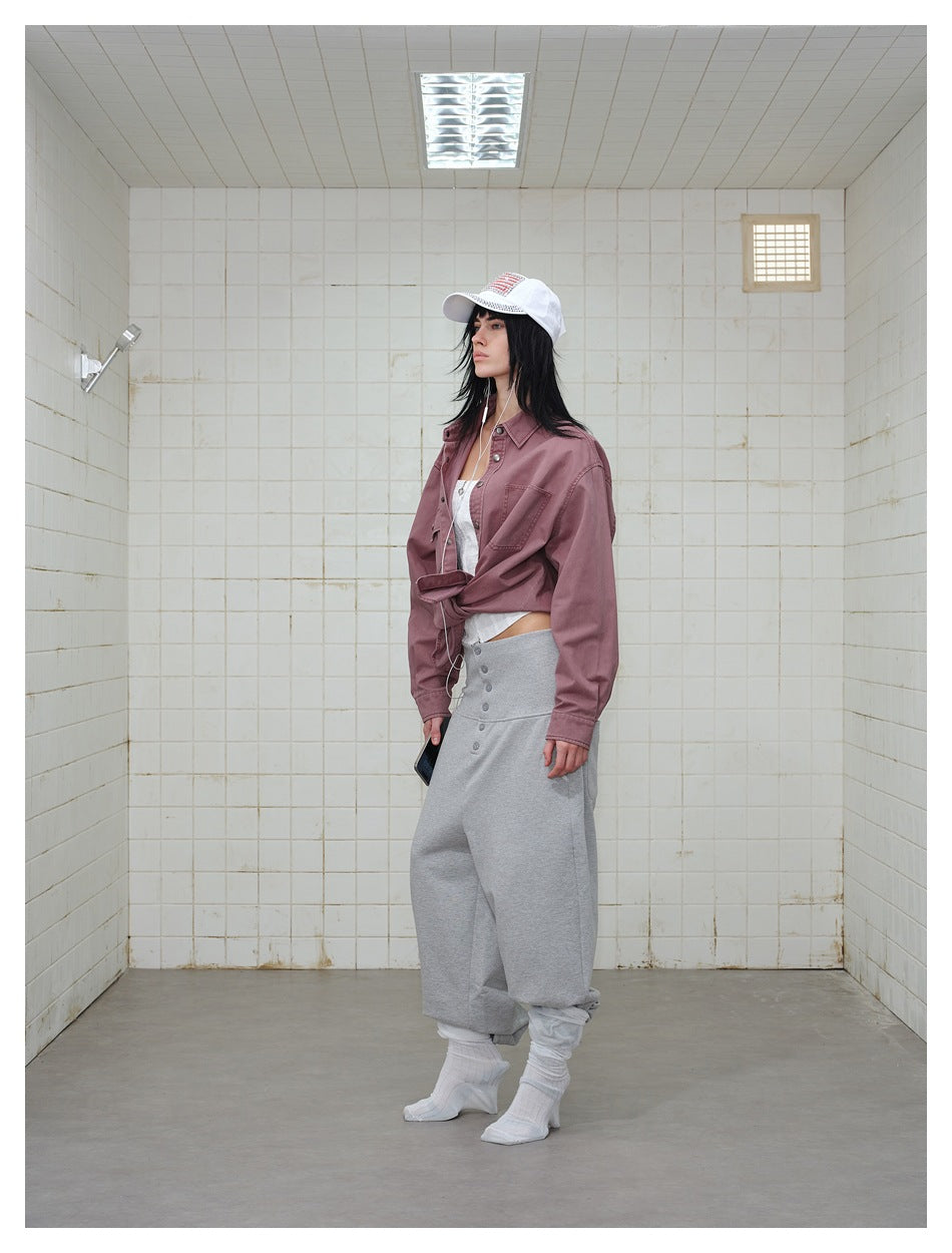 Side view of model in the dusty red shirt tied at the waist with gray wide pants and white cap in a tiled room