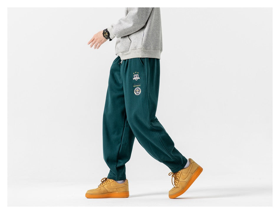 Side view of a model wearing dark teal relaxed-fit joggers with tapered cuffs and small embroidered badges.