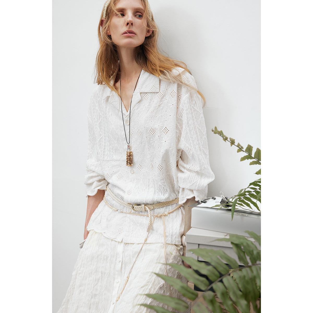 Model wearing a white eyelet button-front blouse with scalloped cuffs, styled with a white skirt and braided belt.