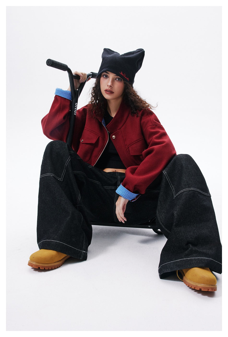 Model seated on a scooter wearing a red jacket with blue checked cuffs, black wide-leg jeans and a black pointed beanie.
