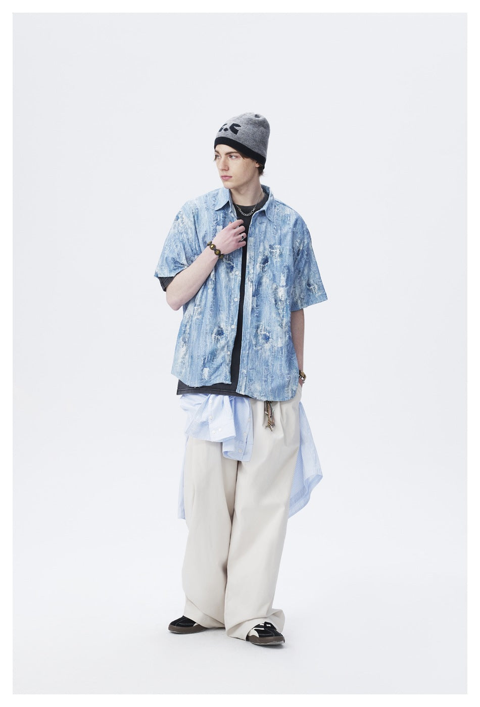 Model in a light blue weathered-print short-sleeve shirt worn open over a dark tee, turned to the side, with beige pants