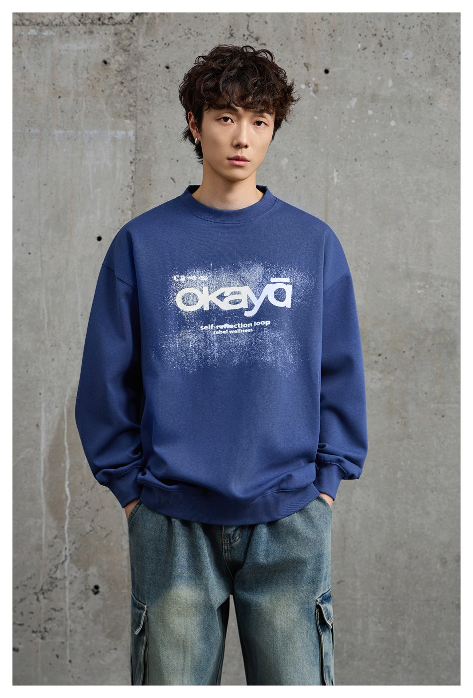 Model wearing a blue crew-neck sweatshirt with white okaya graphic and light-wash cargo jeans against concrete