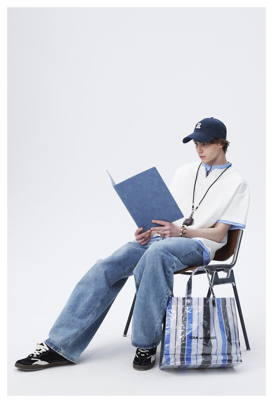 Seated model wearing a white oversized top with blue striped trim, shown with jeans and accessories.