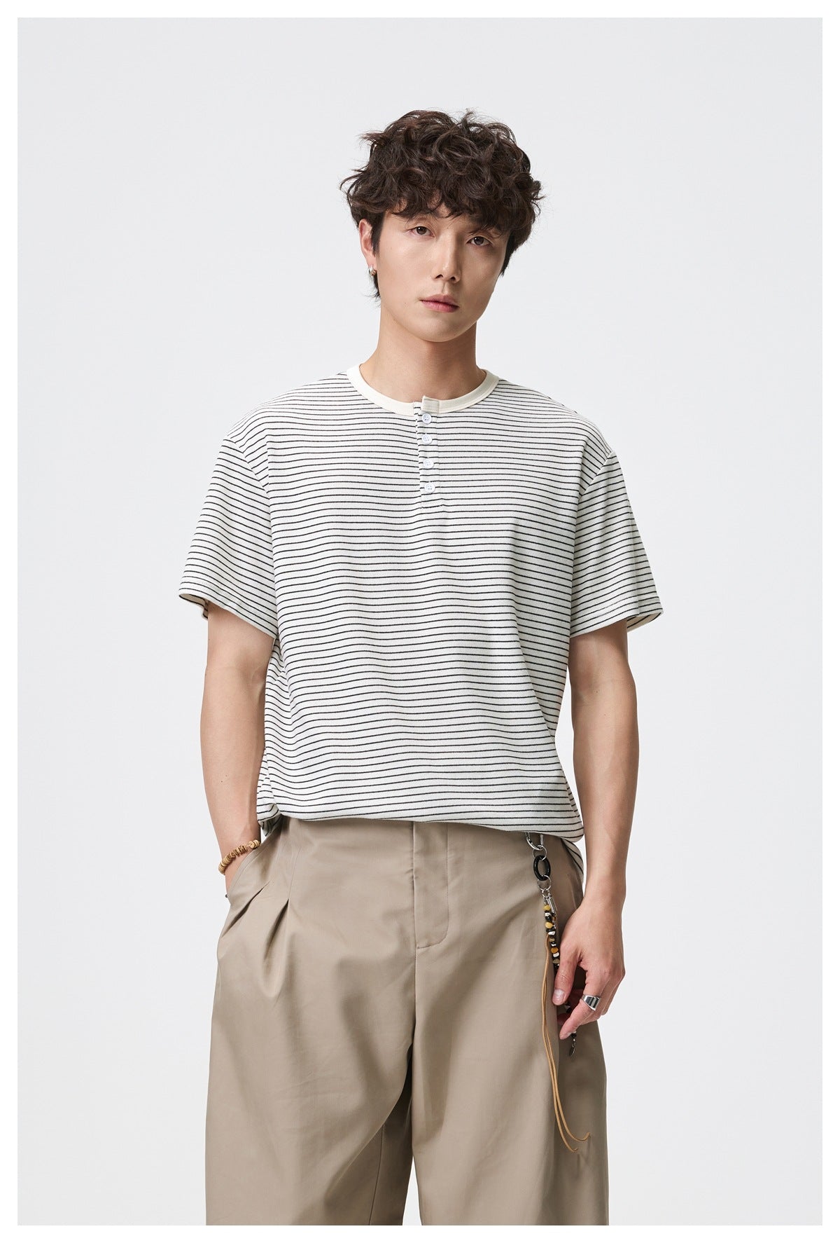 Model wearing an ivory short-sleeve Henley T-shirt with thin black stripes and beige trousers.