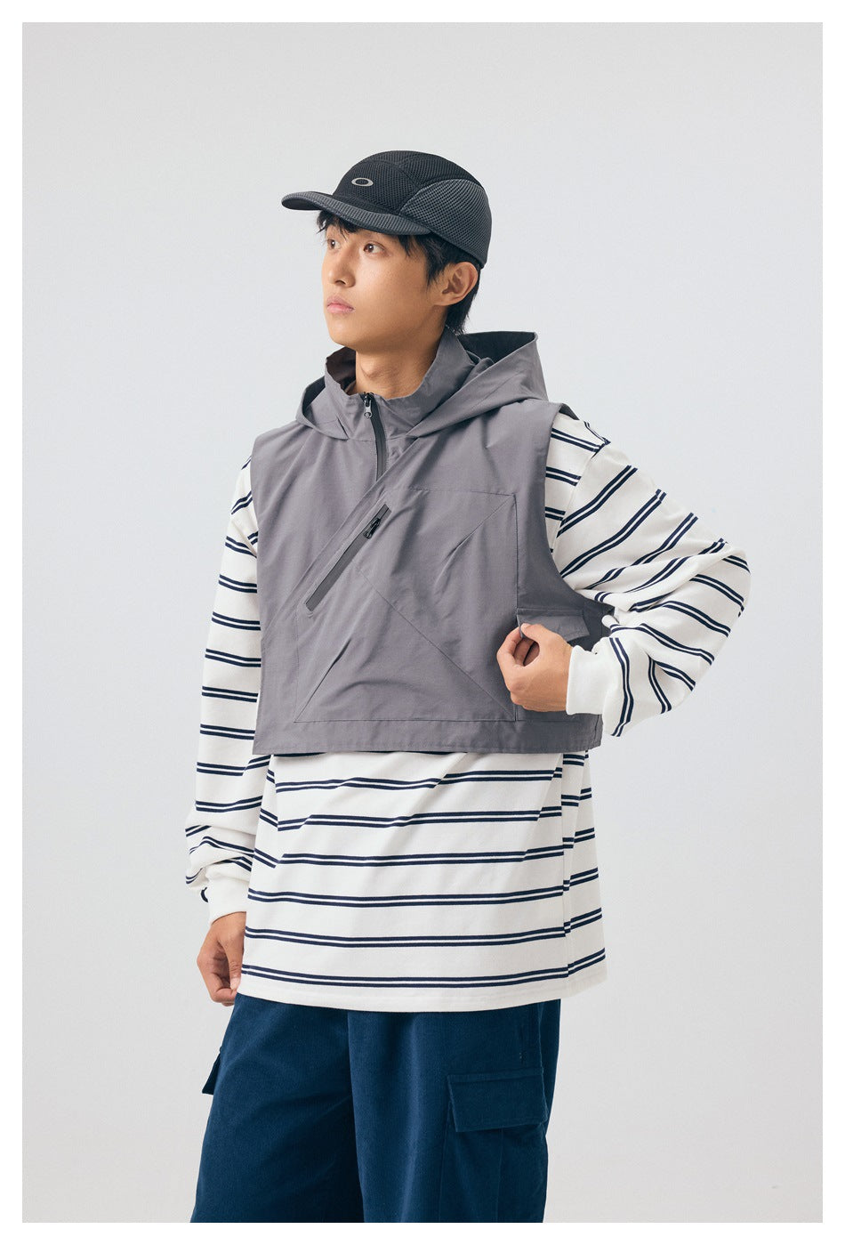 Model wearing a gray cropped hooded vest with a diagonal zip pocket over a white and navy striped shirt.