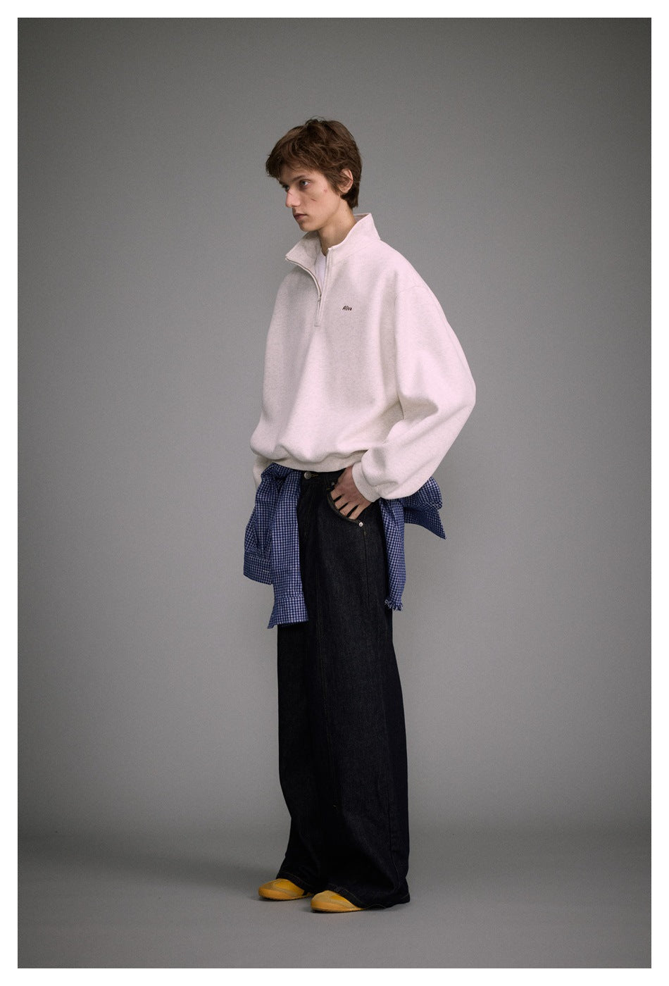 Full-length model wearing a light oatmeal quarter-zip sweatshirt with small chest embroidery and wide dark jeans.
