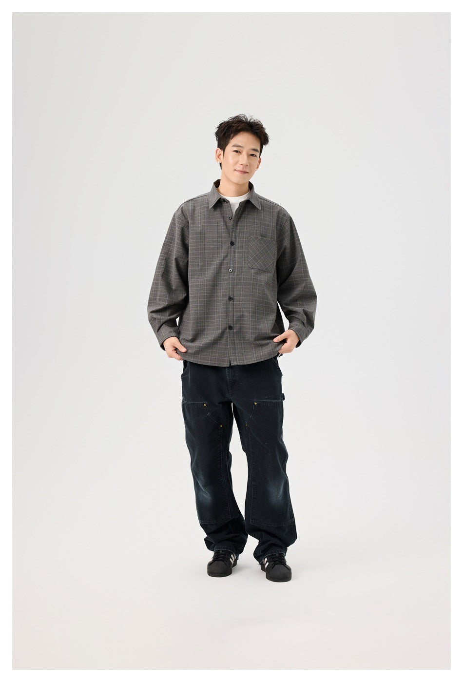 Full-length model wearing a gray plaid button-front overshirt with chest pocket, white tee, loose black pants, and sneakers.